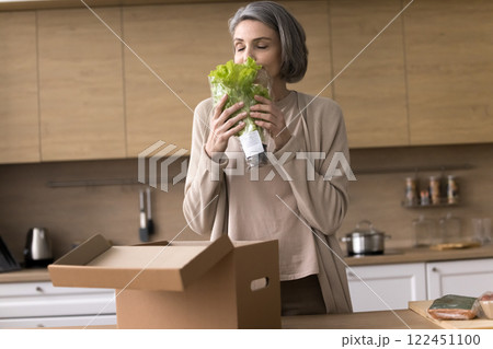 Happy mature woman stand by kitchen table smell fresh salad 122451100