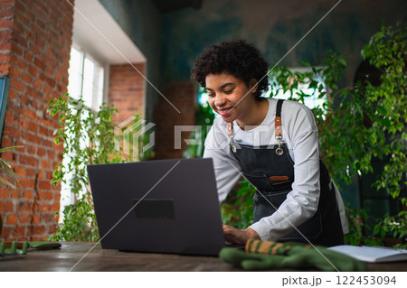 African woman florist gardener working in botanical store using laptop order plants communicating on Internet with customer. Female small business owner working at flower shop. Running of own business African woman florist gardener working in botanical store using laptop order plants communicating on Internet with customer. Female small business owner working at flower shop. Running of own business 122453094