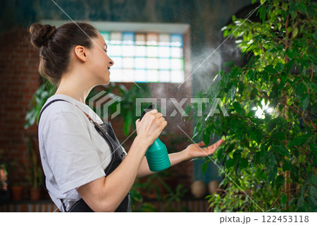 Running of own business. Young woman florist with spray bottle watering fertilizing plants in botanical store. Small business owner working at flower shop smiling surrounded by plants Small business 122453118