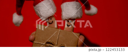 Fingers dressed in Santa-Claus red-white hats. Two happy finger smileys faces holding paper gift box present against red background. Happy family celebrating concept for Christmas or New Years day 122453133