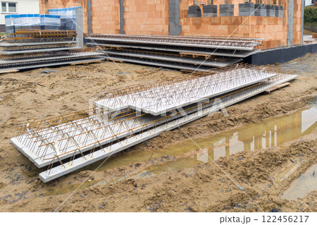 A construction site with stacked reinforced concrete slabs steel supports and a partially built brick structure on sandy ground A construction site with stacked reinforced concrete slabs steel supports and a partially built brick structure on sandy ground 122456217