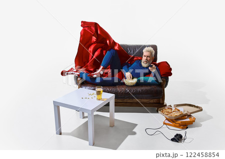 Man in superhero costume with red cape, lounging on couch with beer and snack, holding remote tv controller and watching tv shows against white studio background 122458854