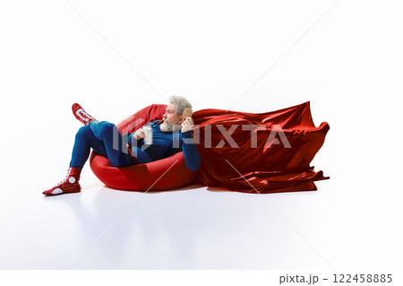 Relaxed grey-haired young man in superhero costume lying on red bean bag, sipping milk and eating cookie against white studio background. 122458885