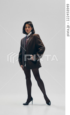 Full-length portrait of a fashionable young woman in black tights and a brown oversized blazer with a tie, confidently standing against minimalistic studio background Full-length portrait of a fashionable young woman in black tights and a brown oversized blazer with a tie, confidently standing against minimalistic studio background 122458949
