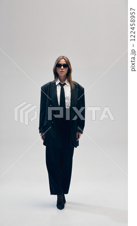 Full-length of confident young businesswoman in black costume, oversized jacket, pants, white shirt and a tie, walking in sunglasses against studio background 122458957