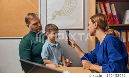 Woman, pediatrician in blue medical coat, examining eye of a boy, child using flashlight. Child sitting on father lap in medical office. Woman, pediatrician in blue medical coat, examining eye of a boy, child using flashlight. Child sitting on father lap in medical office. 122458981