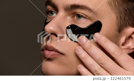 Close up photo of young man with well-kept skin applying moisturized hydrogel black patches against brown studio background. Close up photo of young man with well-kept skin applying moisturized hydrogel black patches against brown studio background. 122461579