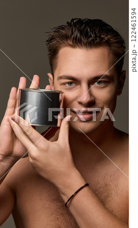 Man smiling while showing blank metallic container near face, tilting head slightly, looking into camera against brown studio background. 122461594