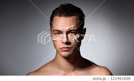 Shirtless man with slicked-back hair staring directly at camera with white pills on forehead and cheeks against dark studio background. Shirtless man with slicked-back hair staring directly at camera with white pills on forehead and cheeks against dark studio background. 122461606