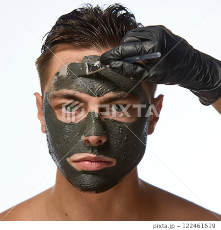 Man with smooth skin looking directly forward, and hand wearing black glove holding brush, mask covering most of face against white studio background. 122461619