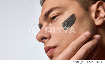 Close up photo of young man with healthy, smooth skin, applying nourishment facial mask against white studio background. 122461631