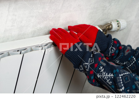 Hands in gloves on a radiator. Central heating radiator at home. A child in woolen gloves warms his hands near the heater Hands in gloves on a radiator. Central heating radiator at home. A child in woolen gloves warms his hands near the heater 122462220