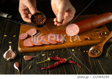 Hands sprinkle colorful spices onto slices of sliced sausage meat, demonstrating culinary skills in a cozy kitchen. The kitchen table setting is conducive to warmth and creativity 122464294