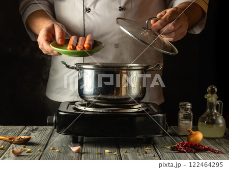 A chef carefully lifts a lid from a pot while holding a plate of sausages, preparing a delicious meal. The cozy kitchen ambiance enhances the cooking experience, blending passion with flavor A chef carefully lifts a lid from a pot while holding a plate of sausages, preparing a delicious meal. The cozy kitchen ambiance enhances the cooking experience, blending passion with flavor 122464295
