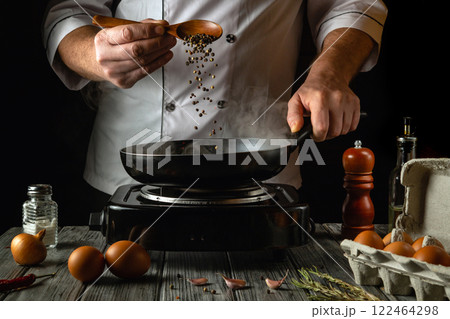 A skilled chef adds fresh peppercorns to a hot pan, releasing aromatic steam. The kitchen is lively with ingredients like eggs, garlic, and herbs, creating a delicious culinary atmosphere A skilled chef adds fresh peppercorns to a hot pan, releasing aromatic steam. The kitchen is lively with ingredients like eggs, garlic, and herbs, creating a delicious culinary atmosphere 122464298