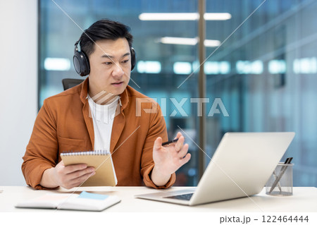 Asian man wearing headphones using a laptop for an online meeting, dressed casually in an office setting. He holds a notepad in one hand and writes with a pen, showing focus and productivity. 122464444