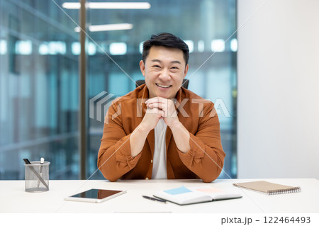 Asian mature businessman seated at a desk, smiling confidently in a professional environment, with office items. 122464493