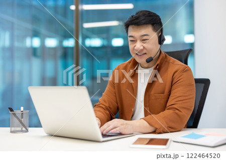 Asian businessman wearing headset working at laptop in modern office. Smiling, focused, multitasking. Smartphone on desk enhances connectivity. Represents professionalism, communication, success. 122464520