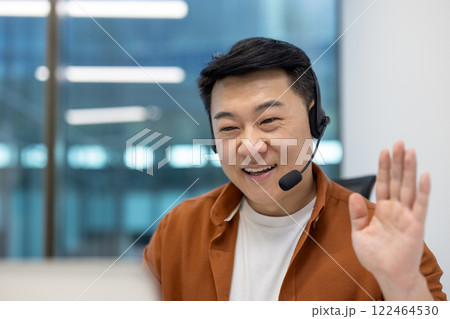 Asian man businessman in video call using headset for online meeting in modern office. Smiling, waving hand, showcasing remote communication, teamwork, technology for workplace connectivity. Asian man businessman in video call using headset for online meeting in modern office. Smiling, waving hand, showcasing remote communication, teamwork, technology for workplace connectivity. 122464530
