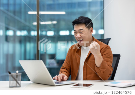 Asian man businessman celebrates success at laptop in modern office setting. Expressions of motivation and achievement highlight success. Enthusiasm evident in smile and fist pump. 122464576