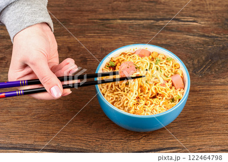 Young woman eating instant noodles with sausages using chopsticks. 122464798