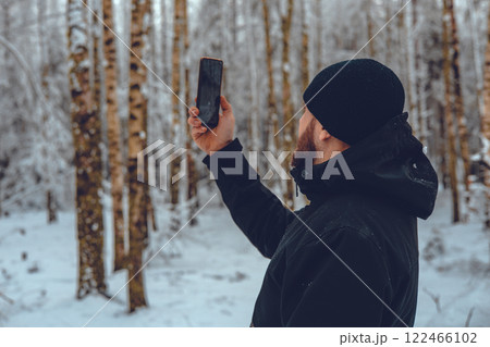 Photographer dressed in warm winter attire captures stunning images with a smartphone amidst a serene snowy forest, fully immersed in the beauty of nature and the peaceful landscape. 122466102