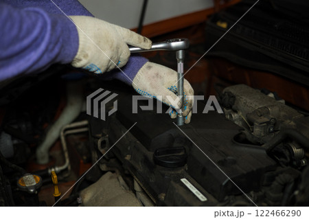 A gloved auto mechanic concentrates on using a ratchet wrench to replace spark plugs in a car engine during routine maintenance at a busy garage workshop. Replacing spark plugs in a car. A gloved auto mechanic concentrates on using a ratchet wrench to replace spark plugs in a car engine during routine maintenance at a busy garage workshop. Replacing spark plugs in a car. 122466290