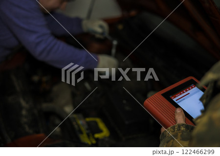Auto mechanics collaborating in a garage, using electronic diagnostic software on a tablet to analyze a vehicle engine, while another mechanic skillfully applying a wrench for repairs. Auto mechanics collaborating in a garage, using electronic diagnostic software on a tablet to analyze a vehicle engine, while another mechanic skillfully applying a wrench for repairs. 122466299