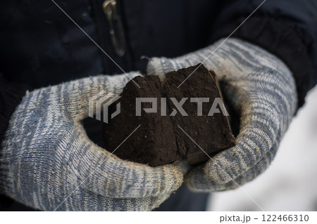Peat briquette for kindling stove in hands. Close-up of farmer's hands in gloves holding peat briquettes - a fuel source for kindling stoves, providing heat on cold winter days in rural areas. Peat briquette for kindling stove in hands. Close-up of farmer's hands in gloves holding peat briquettes - a fuel source for kindling stoves, providing heat on cold winter days in rural areas. 122466310
