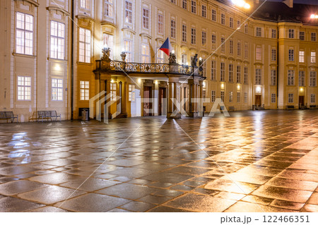 The impressive entrance door to Prague Castle glows under evening lights, with a beautiful balcony adorned by the Czech flag and a reflective courtyard after rain. The impressive entrance door to Prague Castle glows under evening lights, with a beautiful balcony adorned by the Czech flag and a reflective courtyard after rain. 122466351
