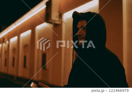 A man in a hood stands on a city street at night with a phone in his hand. The man looks to the side in the light of the city signs at night. Concept of a handsome man on a city street at night. A man in a hood stands on a city street at night with a phone in his hand. The man looks to the side in the light of the city signs at night. Concept of a handsome man on a city street at night. 122466979