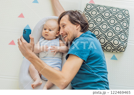 A 3-month-old baby lies safely in a cozy infant cocoon, secured with a special safety belt, while their father lies beside them during a video call. Modern parenting, bonding, and technology concept 122469465