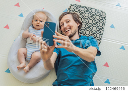 A 3-month-old baby lies safely in a cozy infant cocoon, secured with a special safety belt, while their father lies beside them during a video call. Modern parenting, bonding, and technology concept A 3-month-old baby lies safely in a cozy infant cocoon, secured with a special safety belt, while their father lies beside them during a video call. Modern parenting, bonding, and technology concept 122469466