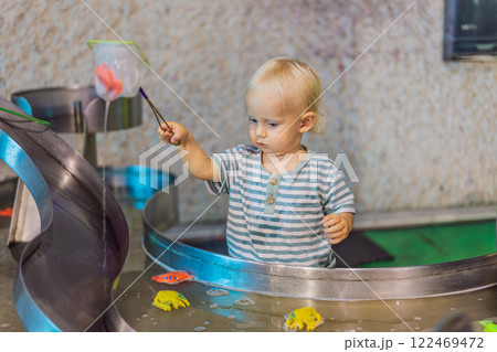 Toddler boy catching toy fish with a net in a vibrant water play area at an entertainment center. A joyful moment of learning, play, and discovery designed to inspire young children Toddler boy catching toy fish with a net in a vibrant water play area at an entertainment center. A joyful moment of learning, play, and discovery designed to inspire young children 122469472