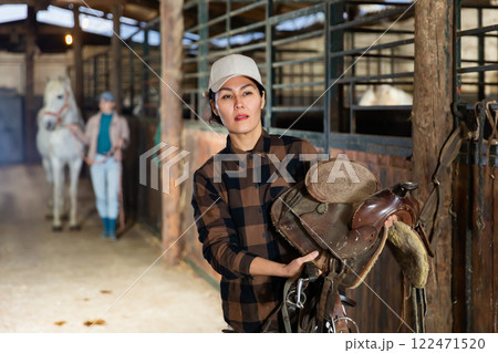 Positive woman carries harness for a horse in stable Positive woman carries harness for a horse in stable 122471520