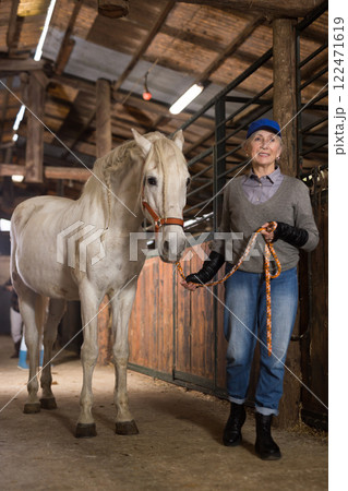 Elderly female farmer holding reins leading white horse through stable 122471619