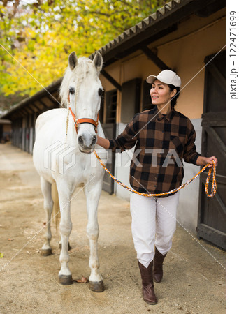 Positive woman jockey leads white horse by the bridle on the street along stable 122471699