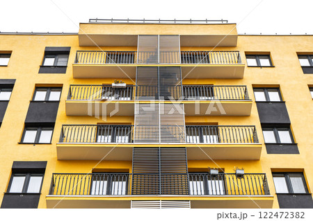 Modern apartment buildings with yellow facade, large windows, and balconies with metal railings, standing against cloudy sky, low angle view Modern apartment buildings with yellow facade, large windows, and balconies with metal railings, standing against cloudy sky, low angle view 122472382