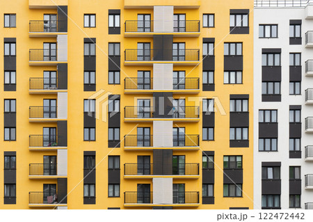 Modern apartment buildings with yellow and white facades, large windows, and balconies with metal railings, against a cloudy sky, front view 122472442