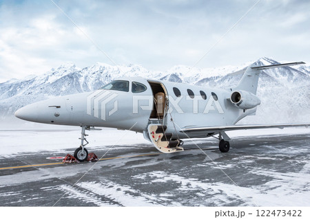 Modern white executive jet plane with an opened gangway door at the winter airport apron on the background of high scenic snow capped mountains Modern white executive jet plane with an opened gangway door at the winter airport apron on the background of high scenic snow capped mountains 122473422