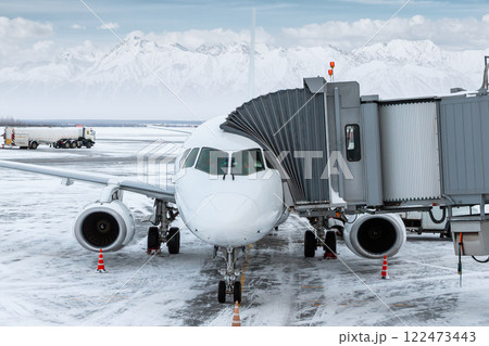 Modern white passenger airliner at the airgate at winter airport apron on the background of high picturesque snow capped mountains Modern white passenger airliner at the airgate at winter airport apron on the background of high picturesque snow capped mountains 122473443