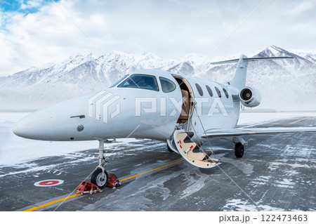Modern white business jet with an opened gangway door at the winter airport apron on the background of high scenic snow capped mountains Modern white business jet with an opened gangway door at the winter airport apron on the background of high scenic snow capped mountains 122473463