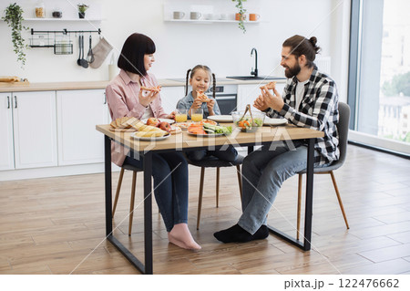 Caucasian family of three having breakfast with pizza and fresh fruits at home. Smiling parents and daughter enjoying meal together. Bright kitchen setting with casual attire and relaxed atmosphere. 122476662