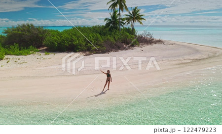 Aerial drone view of a young woman enjoying her vacation on a paradise island, standing on a white sand beach by the turquoise ocean in the maldives 122479223