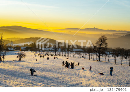 Families and friends enjoy a winter sunset in Prichovice, Czechia, engaging in sledding and playing in the snow while golden hues light up the tranquil landscape. Families and friends enjoy a winter sunset in Prichovice, Czechia, engaging in sledding and playing in the snow while golden hues light up the tranquil landscape. 122479401