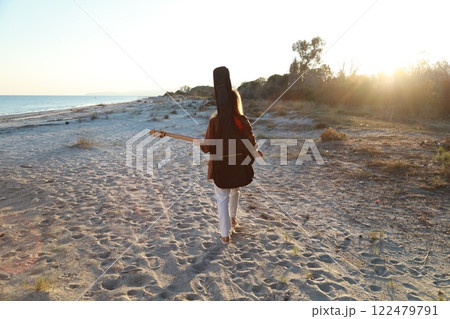 Girl Walks And Plays The Bass Instrument On The Beach Near The Ocean At Sunset Girl Walks And Plays The Bass Instrument On The Beach Near The Ocean At Sunset 122479791