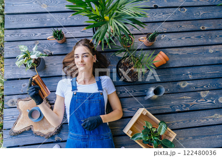 An overhead shot of female gardener lying on wooden terrace holding watering can surround with potted plants, having relax time. 122480036