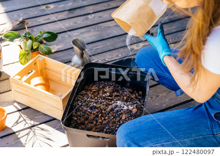 View from above of unrecognizable housewife woman in gloves and casual home clothes mix soil, preparing for plant transplantation on wooden floor terrace outdoor. 122480097