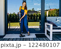 Full height woman housewife using vacuum cleaner on terrace of her house cleaning rug. 122480107