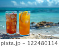 two different cocktails stand in the sand on the edge of the water on a perfect beach in the sand against the backdrop of the blue ocean. 122480114